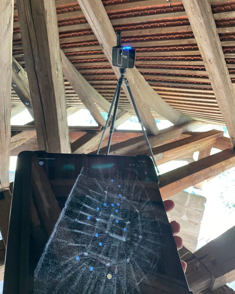 Laserscanner auf einem Stativ in einem historischen Dachwerk, im Vordergrund ein Tablet mit den bereits erfassten Standpunkten